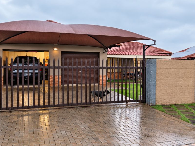 Front view of the property featuring a paved driveway, secure perimeter fencing, and a covered parking area with a shade sail.