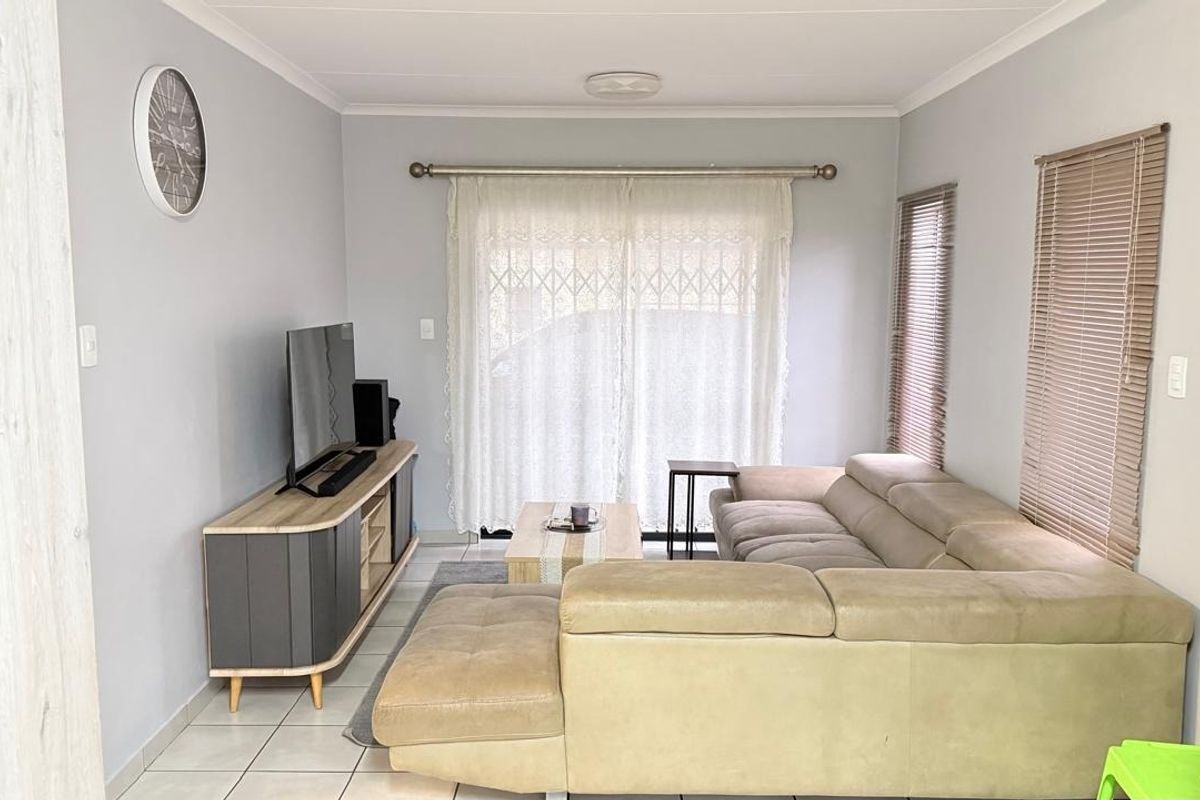 A well-maintained living room featuring neutral wall tones, tiled flooring, and a sliding door with security gate providing access to the exterior.