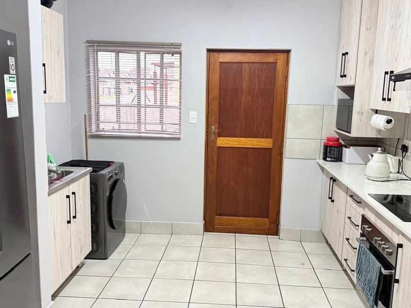 A functional scullery featuring modern light wood cabinetry, stone countertops, integrated appliances, tiled flooring, and a solid timber door leading outdoors.