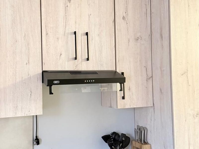 A modern kitchen area featuring light wood-grain cabinetry, a built-in oven, an electric hob, and a black extractor fan with a clean stone countertop.