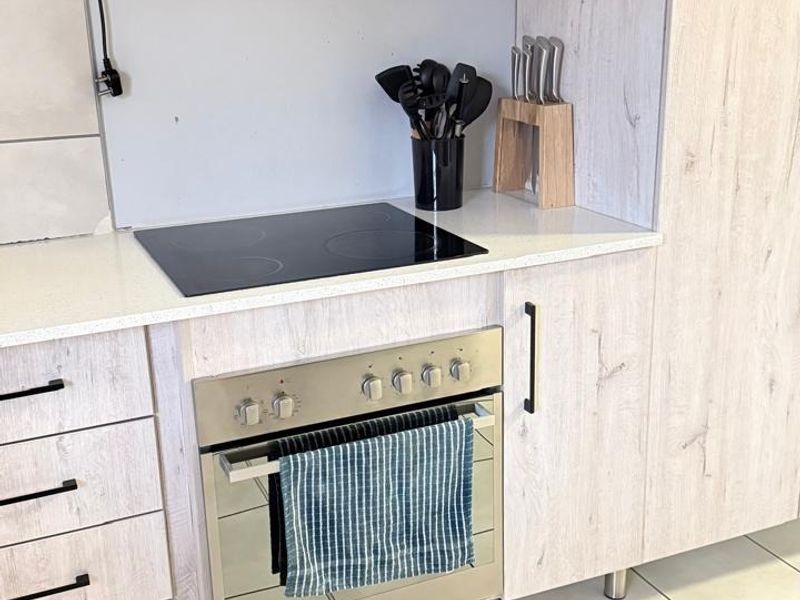 Modern kitchen featuring light wood-grain cabinetry, stone countertops, a built-in oven with electric hob, and an extractor fan, complemented by neutral tiled flooring.
