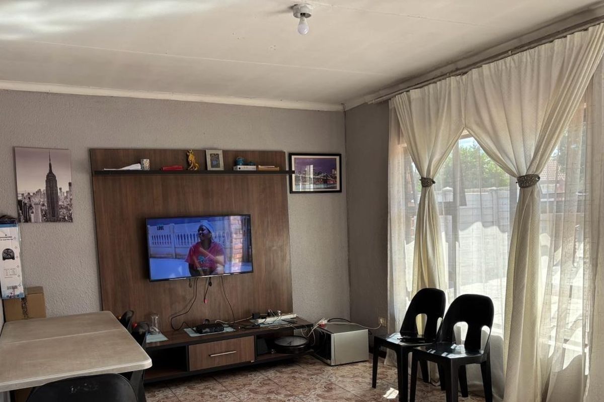 A living room featuring a built-in media unit, tiled flooring, and large windows with curtains, offering a functional space for relaxation.