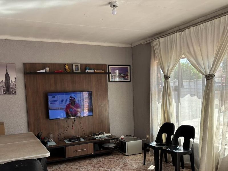 A living room featuring a built-in media unit, tiled flooring, and large windows with curtains, offering a functional space for relaxation.