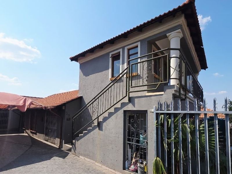 Front exterior view of a two-story house featuring a paved driveway, carport, external staircase leading to an upper balcony, and secure perimeter fencing.