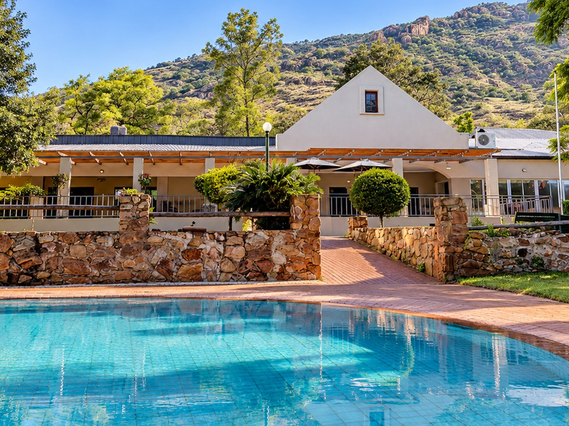 A well-maintained swimming pool area featuring a stone retaining wall, brick paving, and a covered patio overlooking the landscaped garden and mountain backdrop.