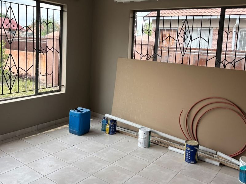 An empty living room space featuring tiled flooring and large windows with security bars, currently undergoing renovation or maintenance work.