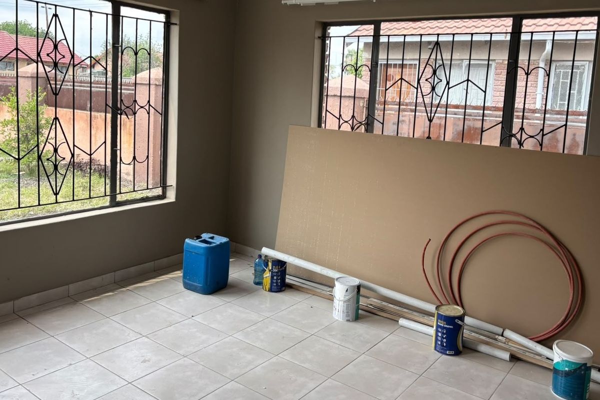 An empty living room space featuring tiled flooring and large windows with security bars, currently undergoing renovation or maintenance work.