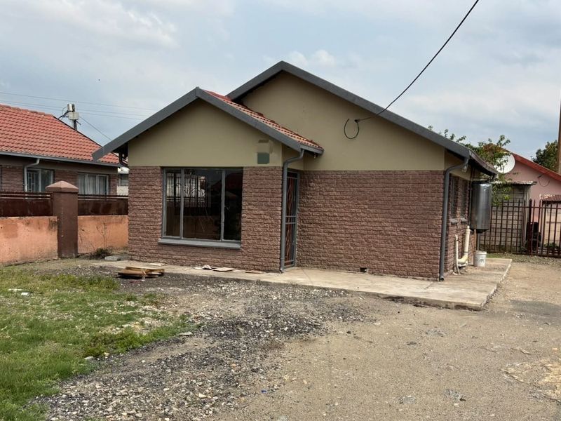 Front view of a single-story brick house featuring a tiled roof, security gate, and an unpaved yard area.