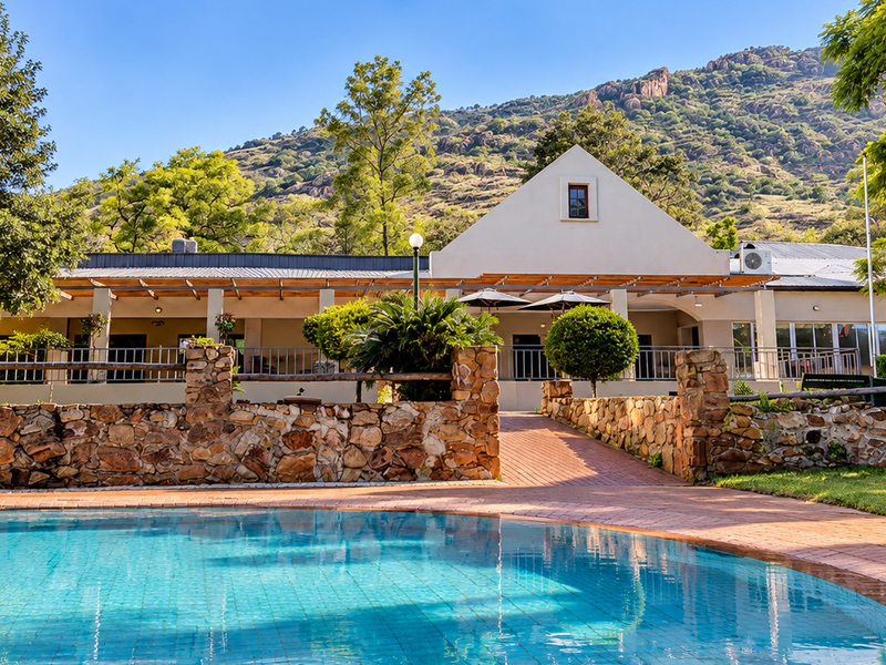 A well-maintained swimming pool area featuring a stone retaining wall, brick paving, and a covered patio set against a scenic mountain backdrop.