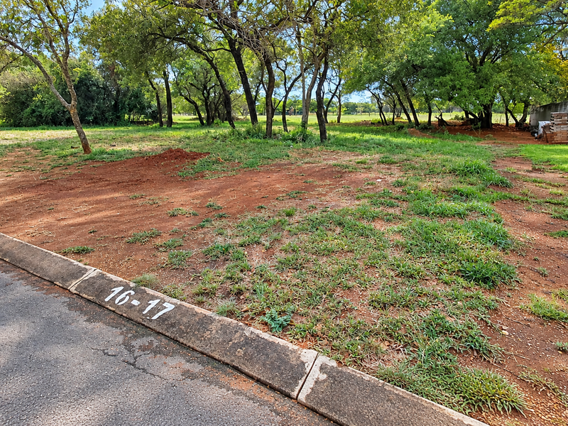A view of a vacant residential plot featuring a concrete curb, asphalt road frontage, and established mature trees.