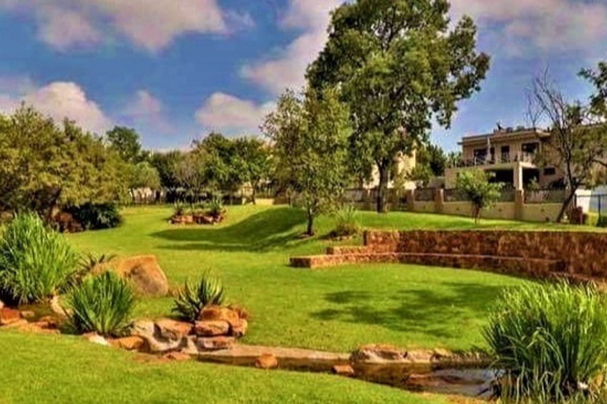 A spacious, well-maintained garden featuring a stone retaining wall, manicured lawn, and a natural stone water feature with the main residence in the background.