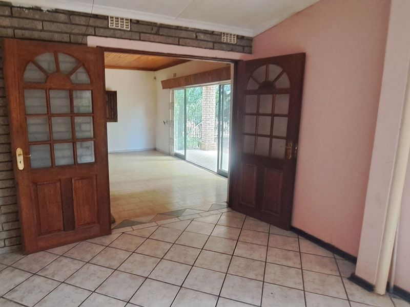 An empty living area featuring tiled floors, an exposed brick feature wall, and double wooden doors leading to an adjacent room with sliding glass access.