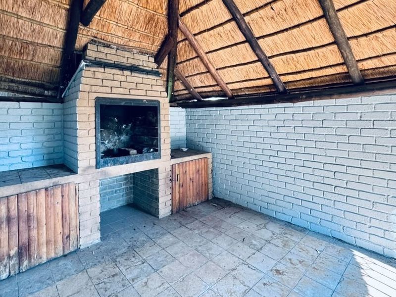 Covered outdoor patio area featuring a built-in brick braai, rustic thatched roof with exposed timber rafters, and tiled flooring.