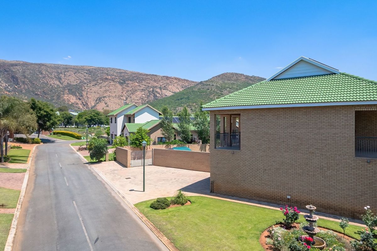 Aerial view of a residential street featuring brick homes with green tiled roofs, paved driveways, and well-maintained garden areas against a mountain backdrop.