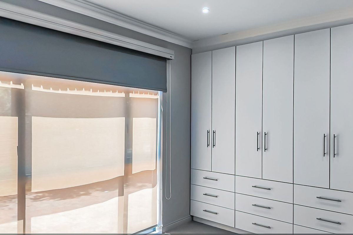 A clean, modern bedroom featuring extensive white built-in cabinetry with integrated drawers and neutral grey walls, complemented by fitted roller blinds.