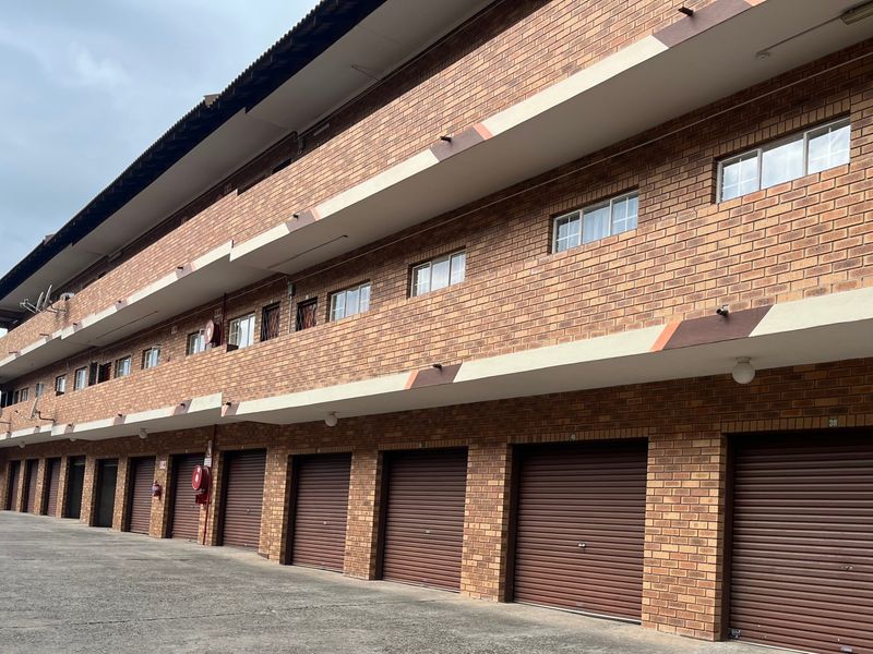 A multi-story apartment building exterior featuring a face brick facade, multiple ground-level roller shutter garages, and a paved driveway.