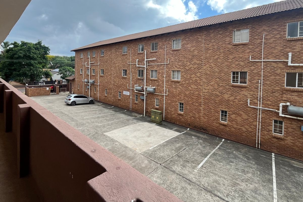 View of the apartment building exterior featuring a brick facade, tiled roof, and a paved parking area with an elevated balcony walkway.