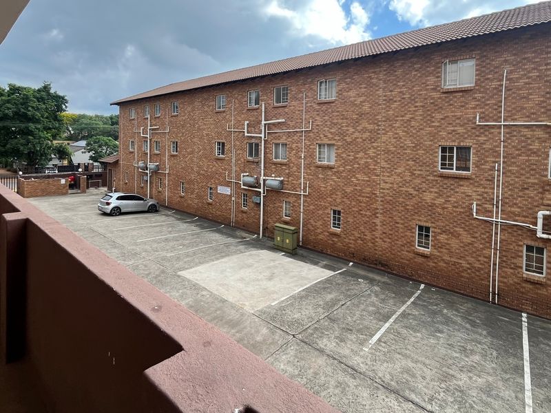 View of the apartment building exterior featuring a brick facade, tiled roof, and a paved parking area with an elevated balcony walkway.