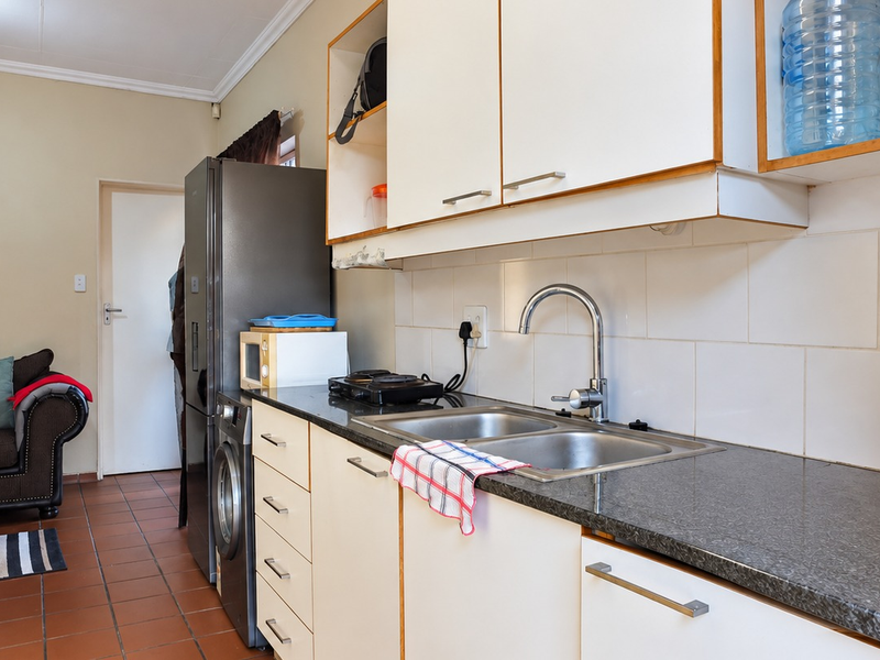 Compact kitchen featuring built-in cabinetry, granite countertops, a double bowl stainless steel sink, and tiled backsplash, with adjacent tiled flooring.