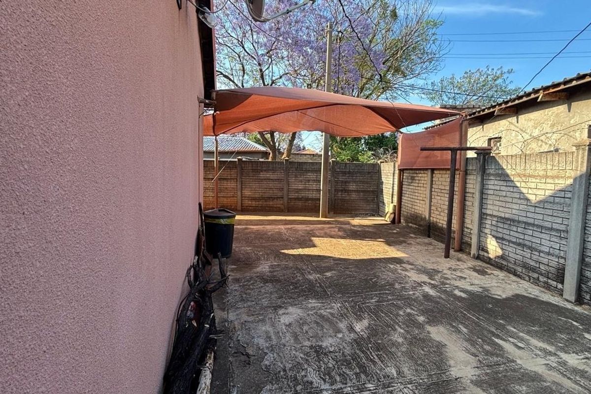 A rear outdoor area featuring concrete paving, a brick boundary wall with security fencing, and a shade sail structure.