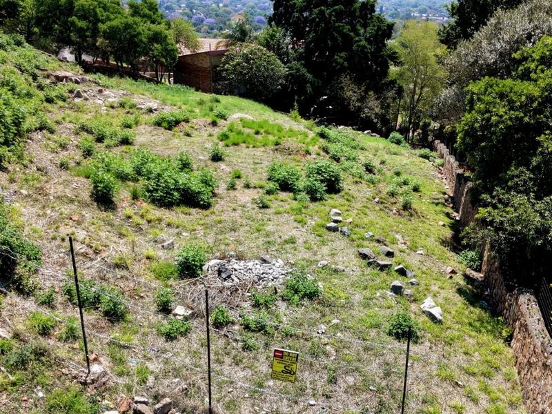 An elevated view of a sloped, vacant residential plot featuring stone retaining walls and perimeter fencing, surrounded by established trees and greenery.