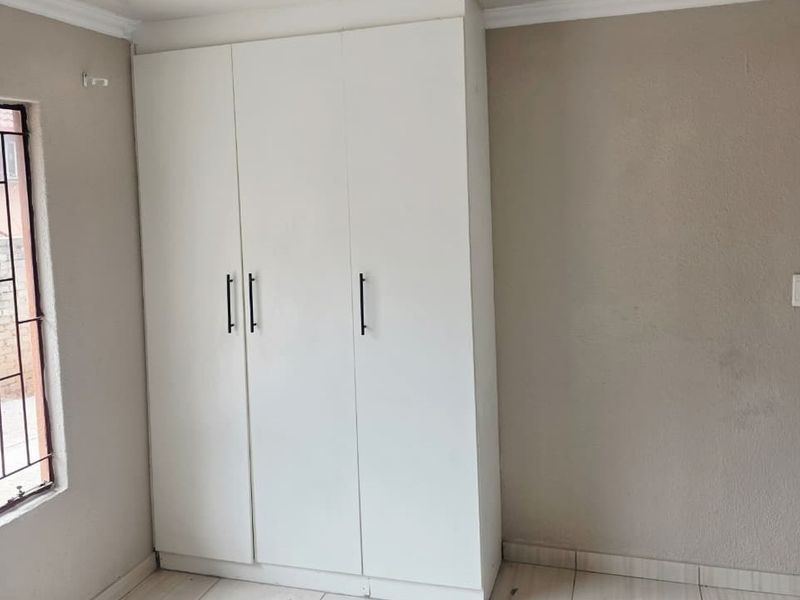 A bedroom featuring built-in white wardrobes, tiled flooring, and security bars on the window, presenting a clean, neutral space ready for personalization.