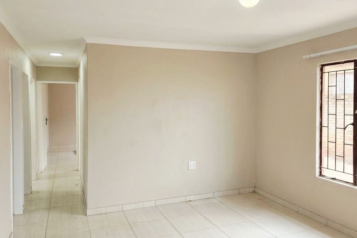 A bright, empty living area featuring neutral wall tones, tiled flooring, and large windows fitted with security bars.