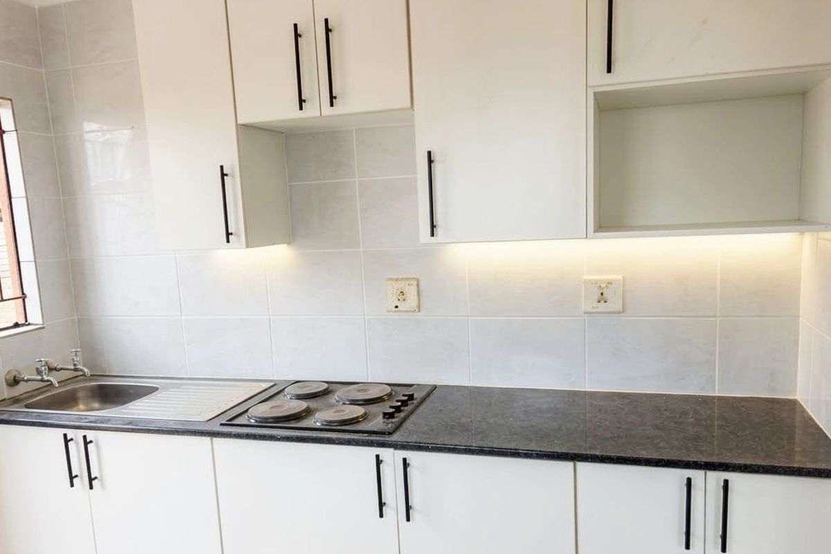 A clean, modern kitchen featuring white cabinetry, dark laminate countertops, a built-in hob, and a stainless steel sink with tiled splashbacks.