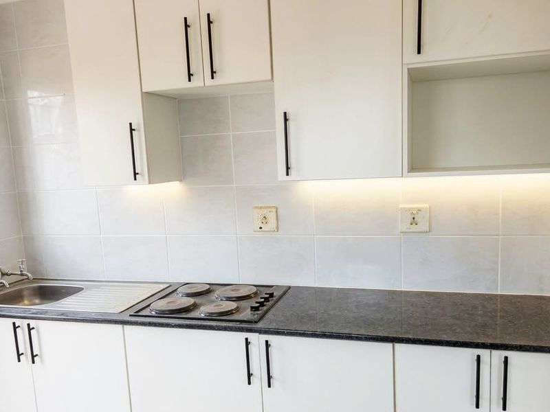 A clean, modern kitchen featuring white cabinetry, dark laminate countertops, a built-in hob, and a stainless steel sink with tiled splashbacks.