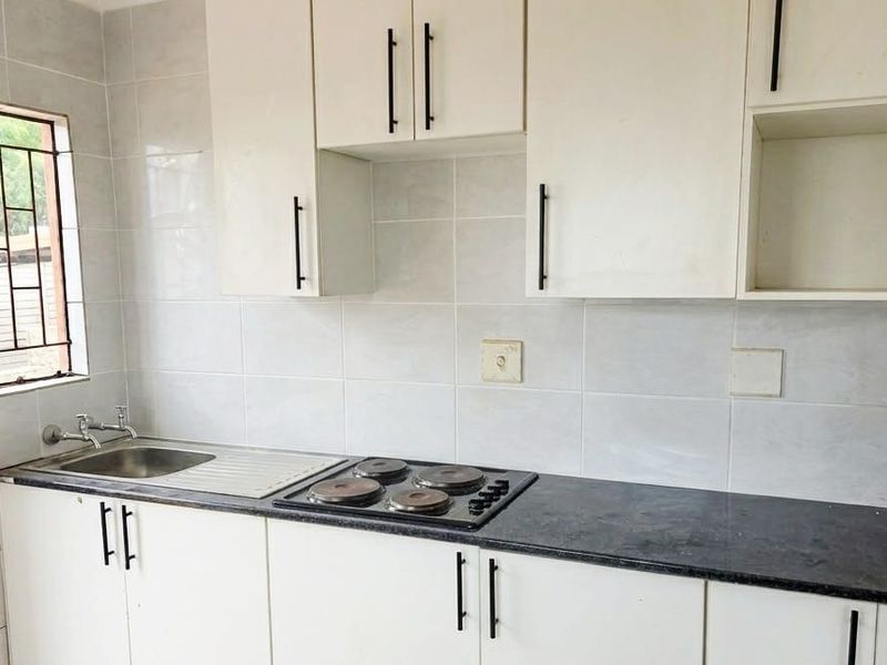 A functional kitchen featuring white cabinetry, a dark countertop, an integrated electric hob, and a stainless steel sink with tiled wall finishes.