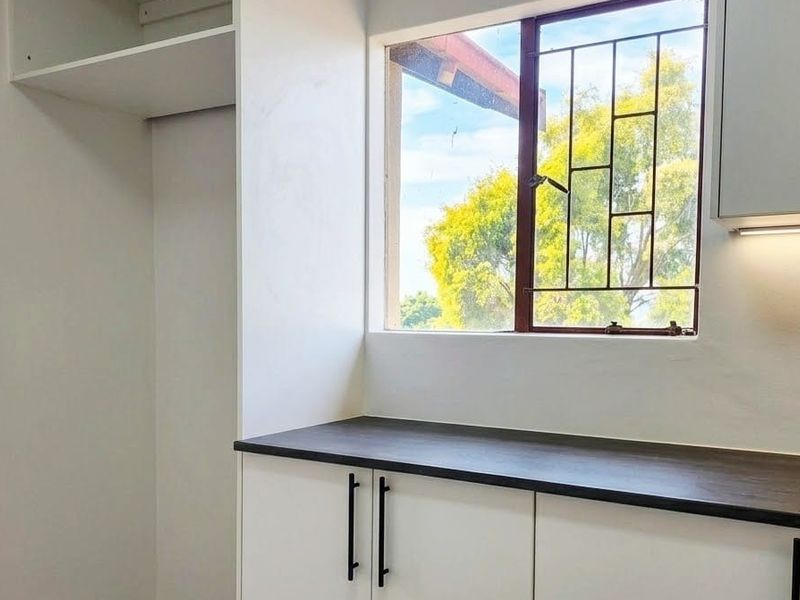 A clean, modern kitchen area featuring white cabinetry, dark countertops, and a window with security bars, offering a bright and functional workspace.