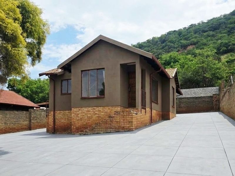 A well-maintained single-story house featuring a brick and plaster facade, tiled roof, and a spacious paved driveway set against a hillside backdrop.