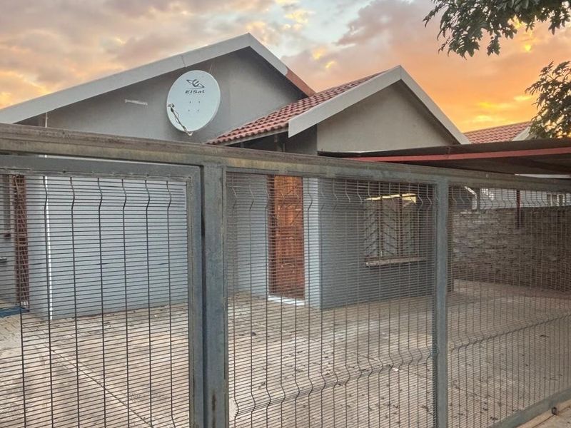 Front view of a residential house featuring a paved driveway, secure metal gate, carport, and tiled roof at sunset.