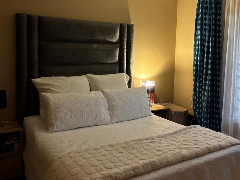 A cozy bedroom featuring a large upholstered headboard, neutral wall tones, and window treatments providing natural light.
