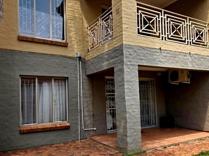Rear exterior view of a multi-story residential building featuring a ground floor patio, upper balcony with metal railings, and security fixtures on windows and doors.