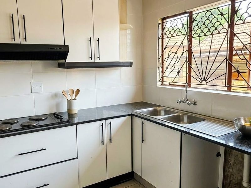 A well-maintained kitchen featuring white cabinetry, dark granite countertops, an integrated electric hob with extractor fan, and a double sink with window view.
