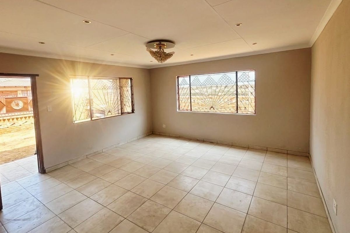 A spacious, empty living room featuring neutral tiled flooring, security burglar bars on windows, and recessed ceiling lighting.