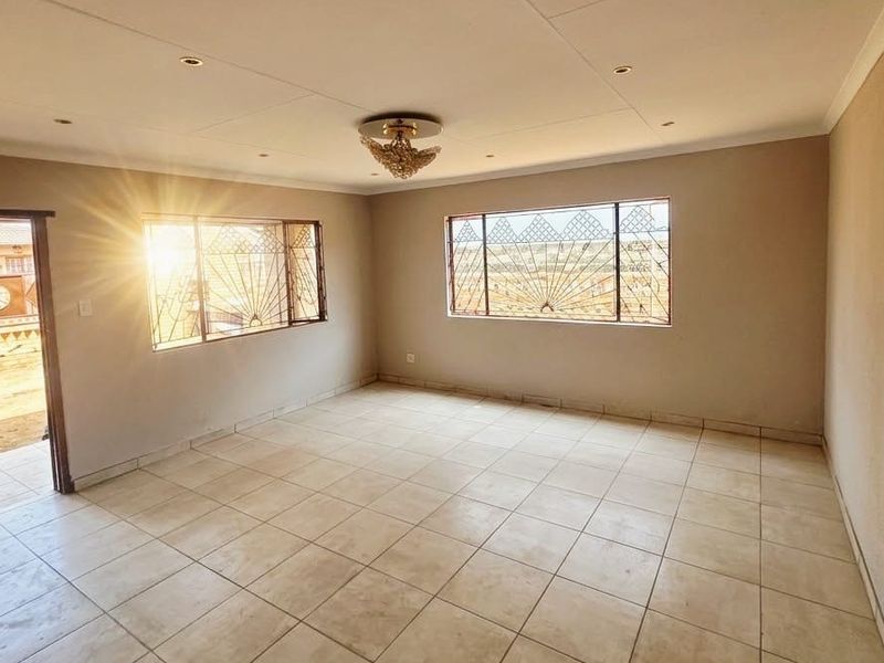 A spacious, empty living room featuring neutral tiled flooring, security burglar bars on windows, and recessed ceiling lighting.