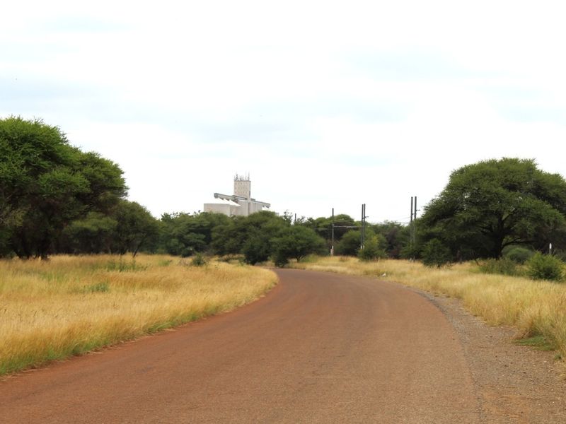 A view of a paved road leading through a rural landscape with surrounding trees and distant industrial infrastructure.