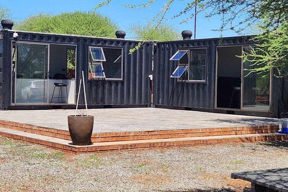 Modern shipping container conversion featuring a spacious brick patio, sliding glass doors, and functional casement windows, set in a rural farm environment.