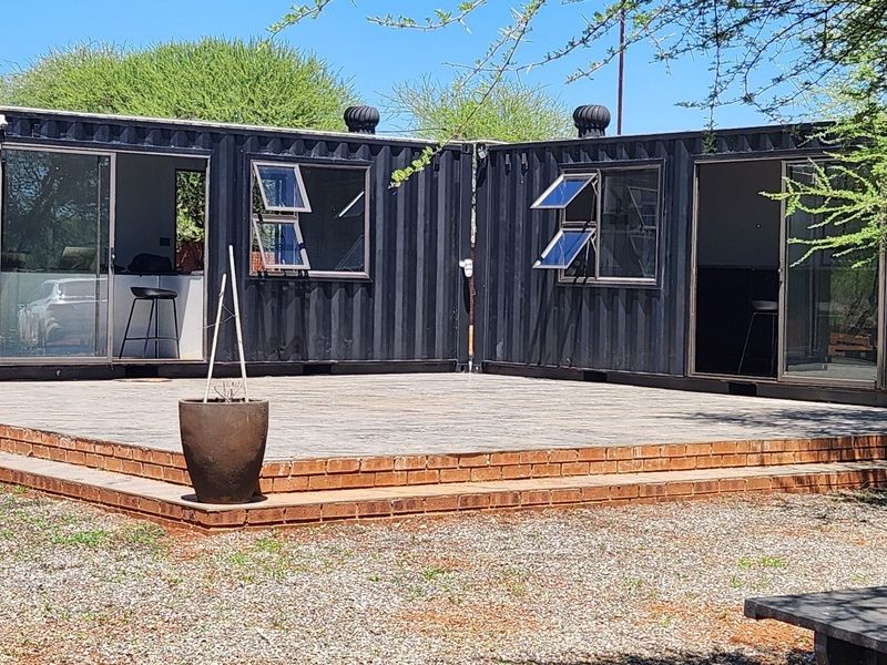 Modern shipping container conversion featuring a spacious brick patio, sliding glass doors, and functional casement windows, set in a rural farm environment.