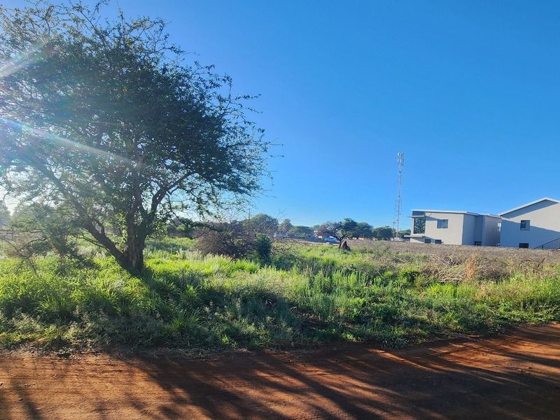 A view of a vacant residential plot featuring natural vegetation and dirt road access, situated in a developing neighborhood.