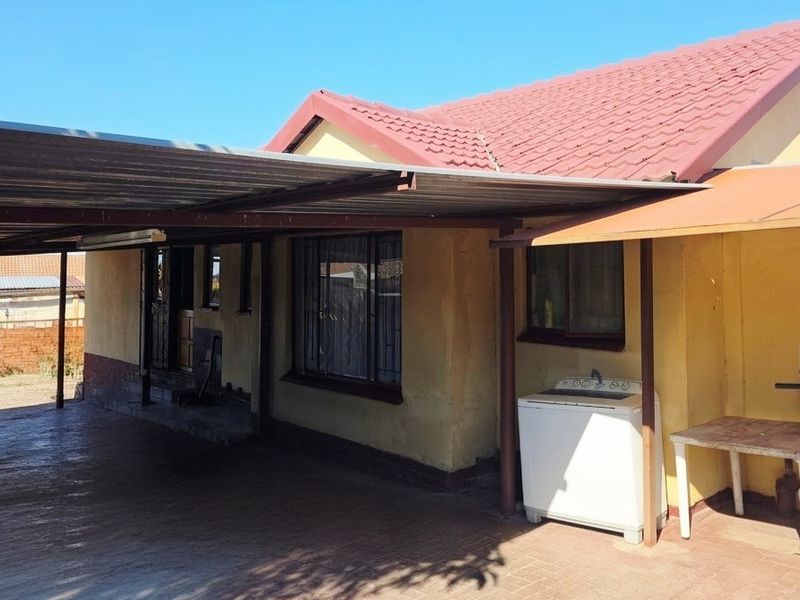 Front exterior view of the house featuring a large carport, paved driveway, and tiled roof, showing signs of weathered exterior wall finishes.
