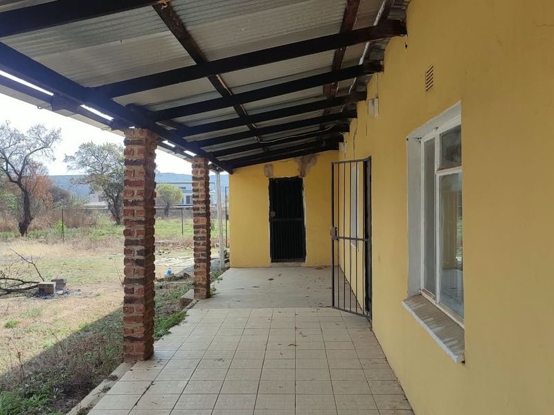 A covered outdoor patio area featuring tiled flooring, exposed brick support pillars, and a corrugated metal roof, overlooking the rear garden space.
