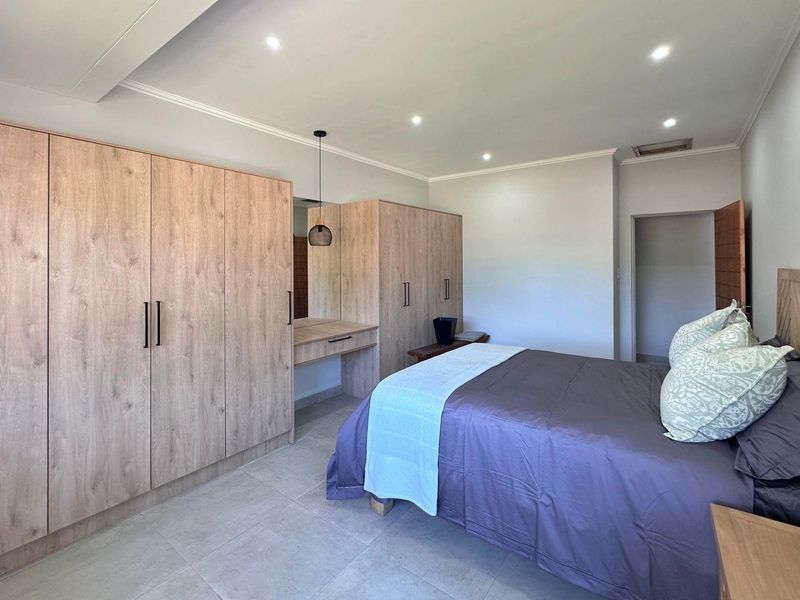 A well-maintained bedroom featuring modern light wood built-in cupboards, a matching vanity, recessed ceiling lighting, and neutral tiled flooring.
