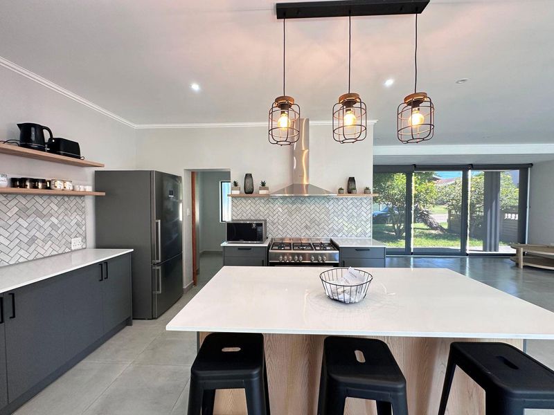 Modern open-plan kitchen featuring a central island, quartz countertops, herringbone backsplash, gas hob, and sleek cabinetry, flowing into a bright living area.
