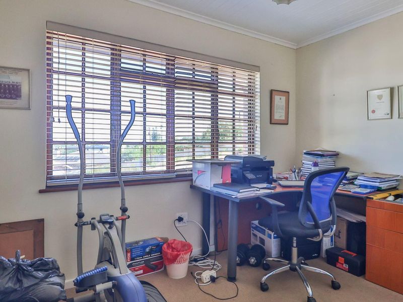 A functional home office space featuring carpeted flooring and large windows with fitted venetian blinds, providing natural light and a view of the exterior.