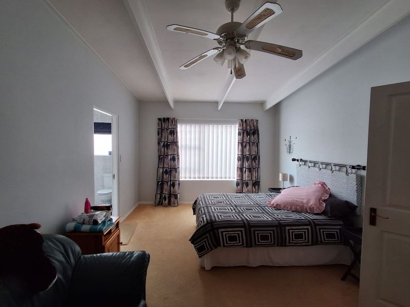A spacious bedroom featuring exposed ceiling beams, a central ceiling fan, and carpeted flooring, with an adjoining en-suite bathroom visible.