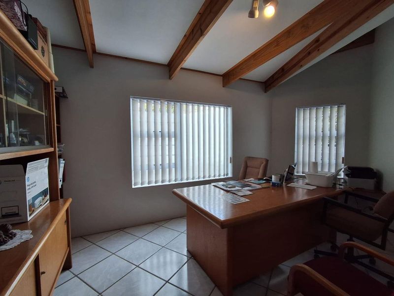 A functional home office featuring exposed timber ceiling beams, tiled flooring, and large windows with vertical blinds, providing a bright workspace.