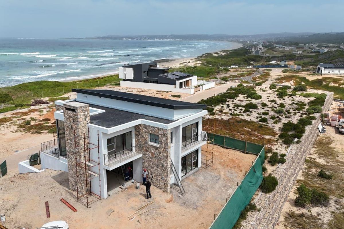 Aerial view of a modern coastal residence under construction, featuring stone cladding, flat roof design, and expansive ocean views.